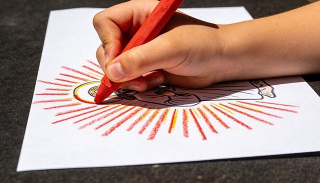 Child coloring sun halo with red crayon close-up, hand holding crayon, outdoor sunlight