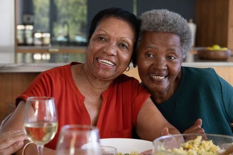 Two african american senior women leaning close smiling over pasta and wine together