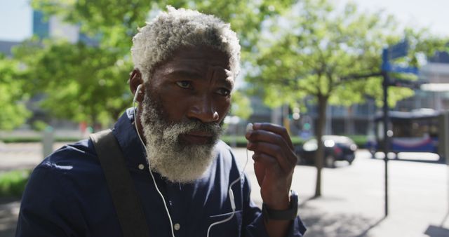 Older man with gray hair and beard listening to music through earbuds while walking in an urban setting with trees in the background. Ideal for topics related to seniors, urban lifestyle, outdoor activities, and modern technology's impact on older generations.