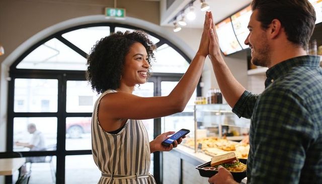 Smiling woman high-fiving man holding smartphone after contactless payment in bakery