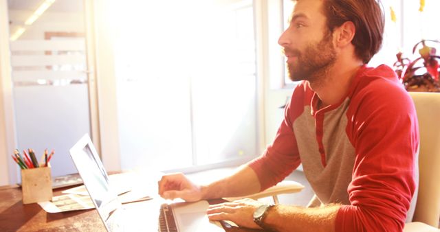 Young man concentrating on work on a laptop in a sunlit office. Ideal for business, technology, and productivity concepts. Can be used by websites, articles, or advertisements focusing on modern work environments, entrepreneurship, and the freelancer lifestyle.