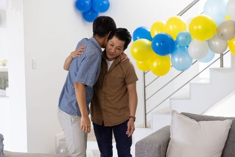 Two adult asian men embracing during home celebration with blue and yellow balloons and cozy living