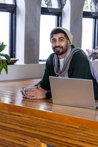 Working remotely at cozy wooden counter with laptop and coffee, smiling man in beanie and scarf