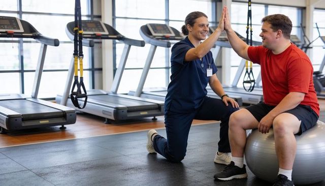Personal trainer high-fiving overweight client on stability ball during gym rehab session