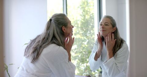 Mature woman with gray hair applying face cream in natural daylight