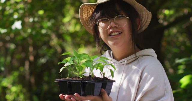 Young woman wearing a hat and glasses, smiling while holding a tray of seedlings in a sunny garden. Ideal for content related to gardening, sustainable living, outdoor hobbies, and lifestyle. Perfect for illustrating themes of nature, growth, and sustainable practices.