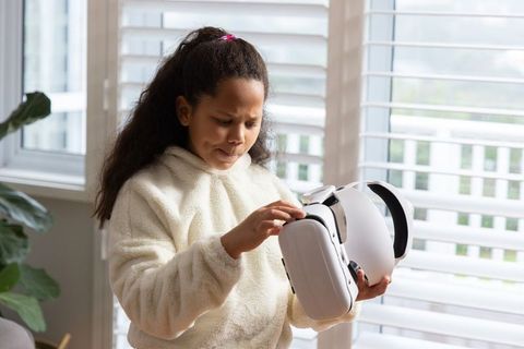 Curious child wearing cream hoodie adjusting white vr headset in bright living room