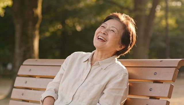 Laughing senior asian woman relaxing on wooden park bench during golden hour