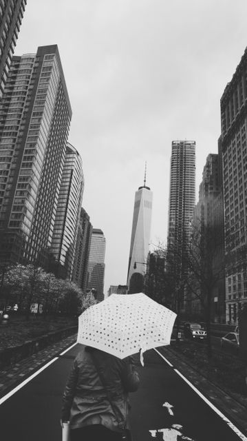 Person walking along city street on a rainy day, holding an umbrella. Surrounded by tall buildings and skyscrapers, providing a modern urban feel. Suitable for use in projects involving city life, travel, weather conditions, solitude, or transportation themes.