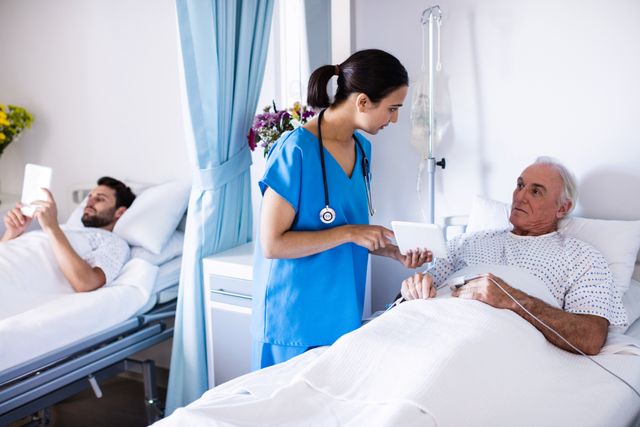 Female doctor discussing treatment options with senior male patient using a digital tablet in a hospital room. Another patient is resting in the background. Useful for healthcare, medical consultation, patient care, and hospital environment themes.
