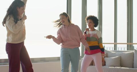 Diverse women singing and dancing in sunlit modern living room holding microphones