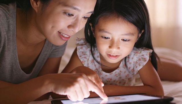 Mother and daughter using tablet in bed at night, cozy bonding and digital learning moment