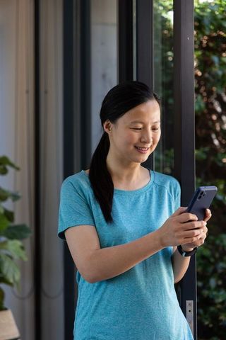 Smiling asian woman checking phone by sliding glass door in blue tee and smartwatch