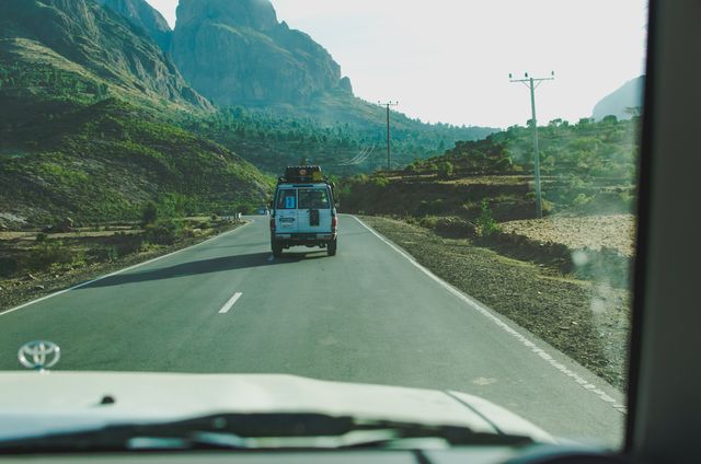 A white van is driving on a remote mountain highway surrounded by lush greenery and rock formations. This image is ideal for use in travel blogs, adventure tourism ads, or outdoor activity promotions, capturing a sense of freedom and exploration.