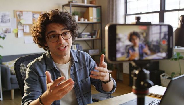 Young vlogger recording casual home office video with gesturing hands and smartphone tripod