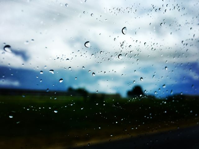 Raindrops on a window creating a scenic, blurry view of the outdoors. Raindrops in focus with background including fields and sky out of focus. Ideal for use in weather-related content, photography displays, or illustrating calm and reflective moods.