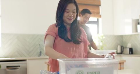 Asian couple sorting recycling in modern kitchen, woman placing bottles into paper bin