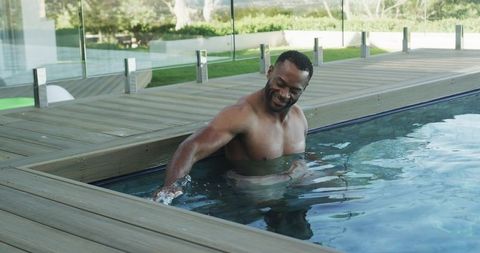 Mature man enjoying poolside dip at modern wooden deck with glass railing