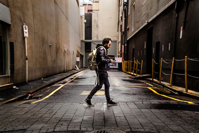 A young adult man with a backpack is walking in a narrow city alleyway while checking his phone. This urban scene could be used in contexts related to navigation, travel, exploration, city life, or technology usage in daily life.