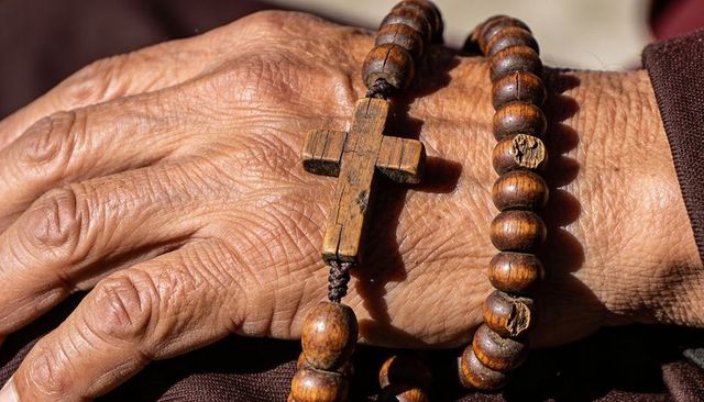 Close-up elderly hand resting with wooden rosary beads and worn cross on burgundy sleeve