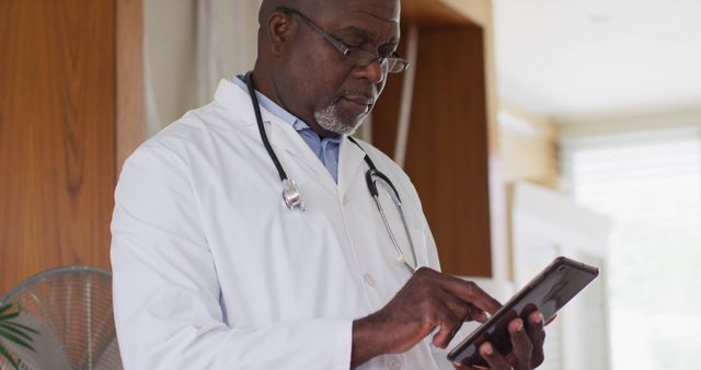 This image depicts an afro-american male doctor in a clinic setting checking a digital tablet. He is dressed in a white lab coat with a stethoscope around his neck, conveying professionalism and expertise. This image could be used for healthcare technology promotion, medical services advertising, educational materials, telemedicine platforms, and doctor-patient interaction visuals.