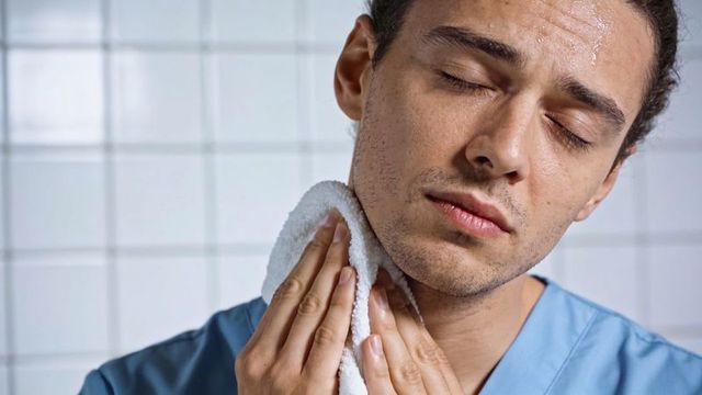 Male clinician pressing towel to neck sweating in tiled room showing fatigue and relief