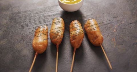 Displaying four golden corn dogs on wooden skewers with mustard on dark slate close-ups