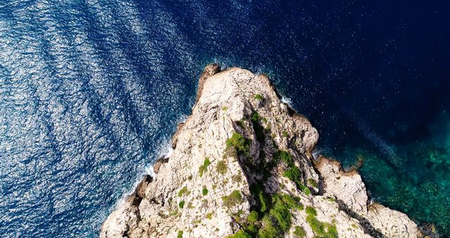 Aerial view captures the rugged beauty of a rocky cliff jutting into the shimmering blue sea. The contrast between the craggy landscape and the deep hues of the ocean emphasizes nature's diverse textures and colors.