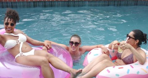 Three women lounging on pink and donut inflatable rings in sunny backyard pool