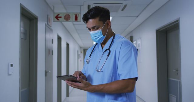 Biracial male doctor wearing face mask standing in hospital corridor using tablet. medicine, health and healthcare services during coronavirus covid 19 pandemic.