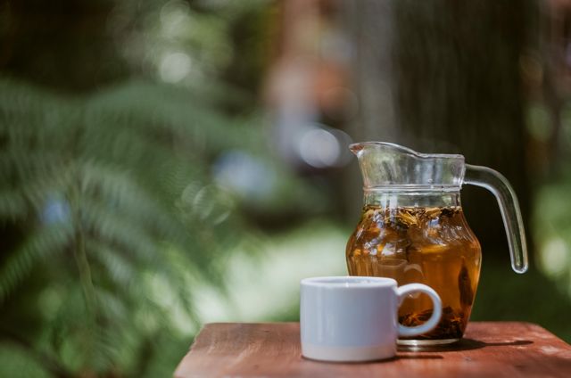 Herbal tea in a glass pitcher with a white mug resting on a wooden table against a forest background. Sunlight filters through the trees, creating a serene and calming atmosphere. Useful for themes related to relaxation, nature, eco-friendly living, and healthy eating.