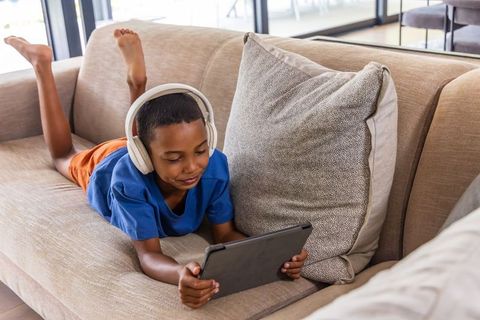 African american boy lying on sofa listening with headphones using tablet in sunlit room