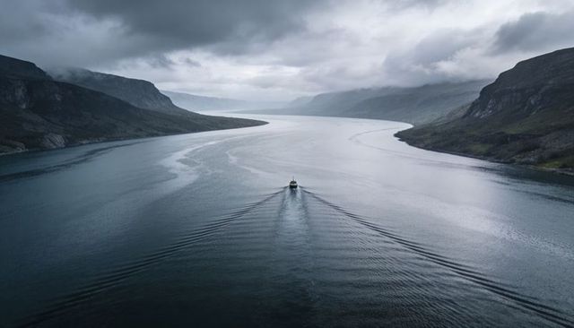 Solitary motorboat cutting calm fjord water with twin v-shaped wakes under moody clouds