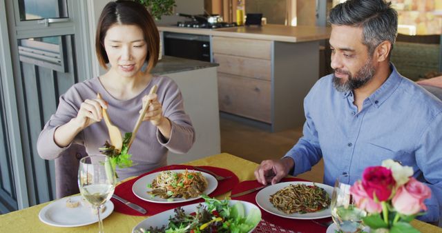 Couple sitting at a modern dining table and enjoying a healthy lunch consisting of salad and noodles. They are seated near a well-lit kitchen, with wine glasses and a floral centerpiece on the table. Ideal for content focusing on healthy eating, relationship bonding, home dining experiences, and lifestyle themes.