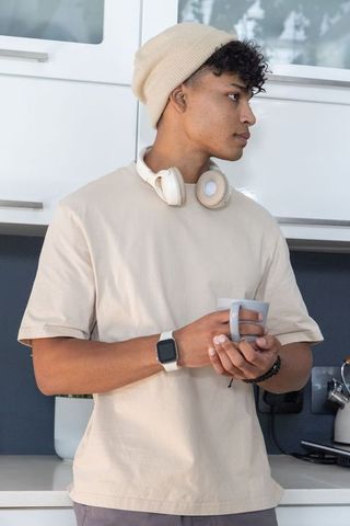 African man standing in kitchen holding mug wearing beanie and headphones, smartwatch