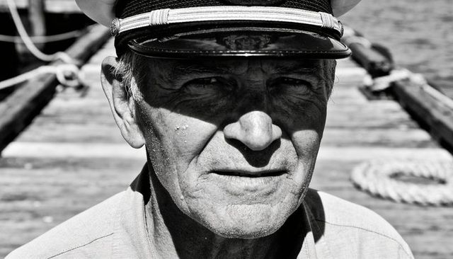 Weathered sea captain squinting on sunlit dock pier wearing peaked nautical cap monochrome close-up