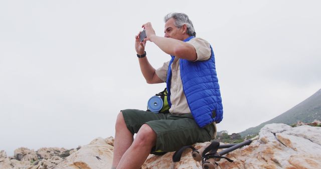 Senior man sitting on rocky terrain taking photos with his camera during a hiking trip in the mountains. Suitable for use in content related to travel, outdoor adventures, hiking tips for seniors, active lifestyle promotion, and encouraging natural photography hobbies.