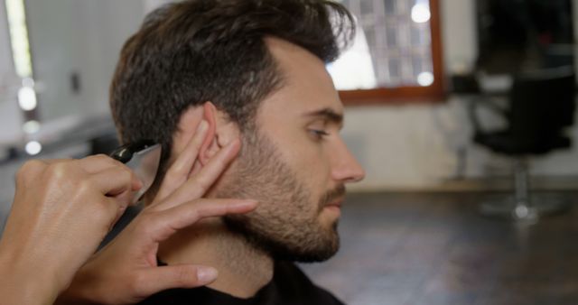 Barber trimming man's hair with clipper, side view focusing on detailing around ear. Ideal for depicting male grooming, barbershop services, hairdressing, and stylist precision in action.
