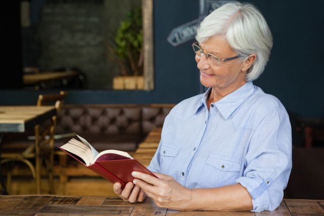 Smiling senior woman reading book while sitting by table at cafe shop