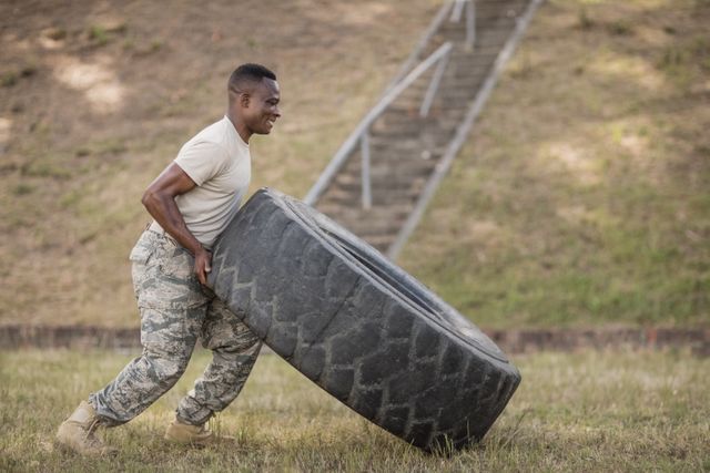 Soldier flipping a large tire outdoors during boot camp training. Ideal for use in articles or advertisements related to military training, physical fitness, strength training, and discipline. Can also be used in motivational contexts or fitness programs.