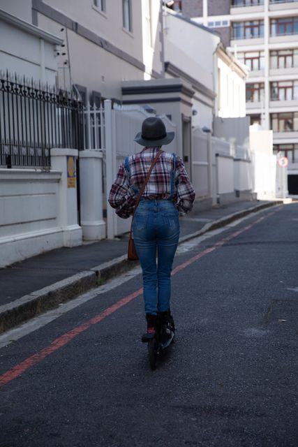 Rear view of biracial alternative woman with short blonde hair, hat and denim dungarees out and about in the city on a sunny day, riding electric scooter. Urban independent woman on the go.