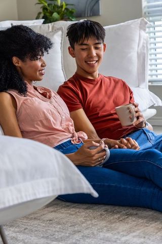 Young couple sitting on floor holding mugs, smiling and chatting in cozy sunlit living room