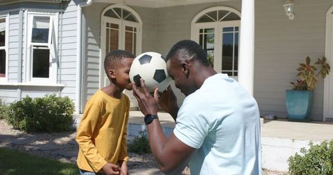 African american father and son bonding pressing soccer ball between foreheads on lawn
