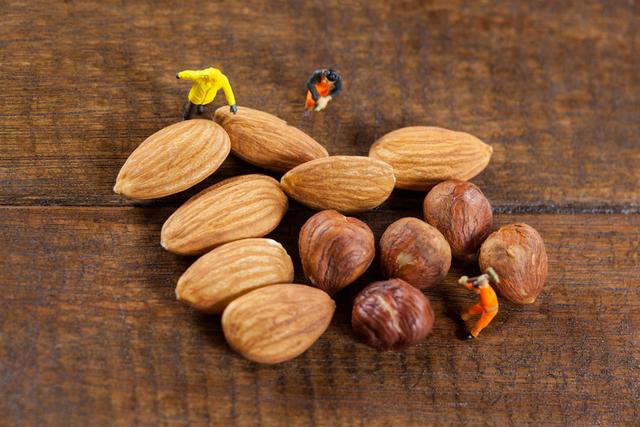 This image depicts miniature workers interacting with almonds and nuts on a wooden surface, creating a playful and creative scene. Ideal for use in advertisements, blogs, or articles related to healthy eating, nutrition, teamwork, and creative concepts. It can also be used in marketing materials for food products or as a fun visual in presentations and social media posts.