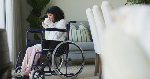 Young woman seated in a wheelchair, sipping coffee in a bright, cozy living room. Suitable for illustrating articles about accessibility, home relaxation, daily routines for individuals with disabilities, or indoor comfort.