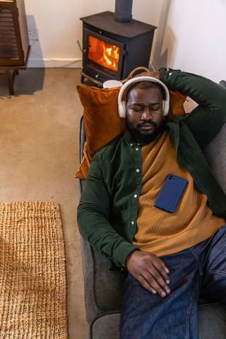 Relaxing african american man listening with headphones on sofa beside glowing wood stove