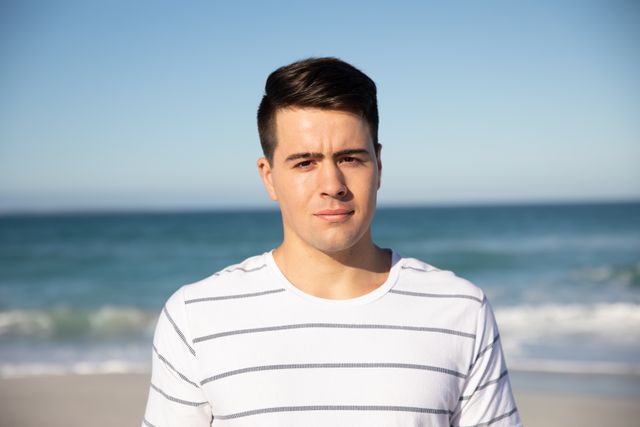 Young Caucasian man standing on beach with ocean in background, wearing striped shirt. Ideal for travel brochures, summer vacation promotions, lifestyle blogs, and advertisements focusing on leisure and relaxation.