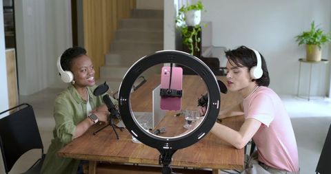 Diverse co-hosts recording podcast at home table with ring light, pink phone, studio gear