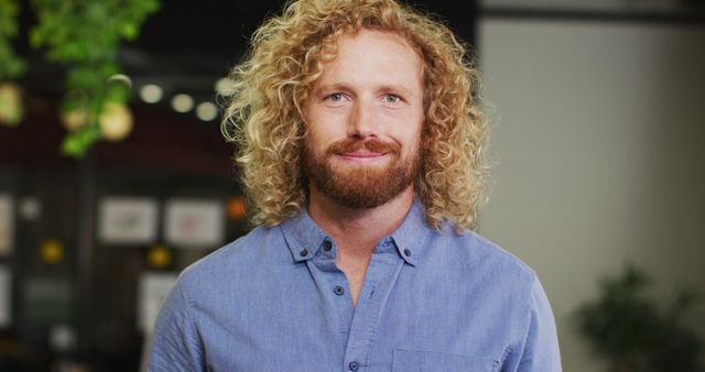 Young man with curly hair and beard smiling confidently indoors while wearing a casual blue shirt. Ideal for use in professional headshots, marketing materials, website banners, or stock photos related to lifestyle, work, confidence, or personal branding.