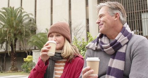 Senior couple sipping coffee on urban bench wearing scarves and knitwear, smiling warmly